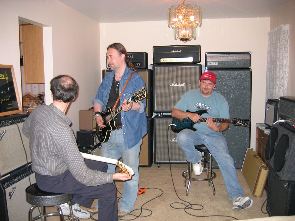 Brian, John, and Ken sit around a room with their respective guitars plugged into a series of large amplifiers